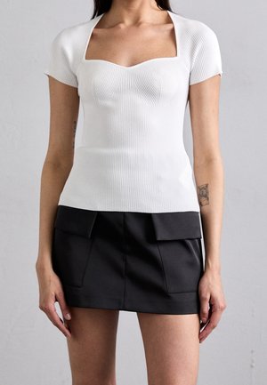 Frau trägt ein weißes, geripptes T-Shirt mit kurzen Ärmeln und quadratischem Ausschnitt sowie einen schwarzen Minirock mit vorderen Klappen-Taschen. Ein Tattoo ist am linken Arm sichtbar.