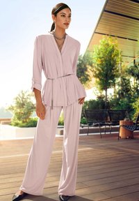 Helles rosa Bluse und weite Hose aus glattem Stoff, akzentuiert mit einem silbernen Gürtel. Das Model trägt schwarze spitze Schuhe und gestaffelten silbernen Schmuck.