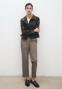 Žena stojící proti bílé zdi, oblečená v černé kožené bundě, bílé blůze, hnědých elegantních kalhotách a černých mokasínách.