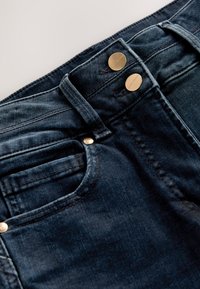 Tmavomodré denimové džínsy zblízka zobrazujú dvojité kovové gombíky, opaskové očká, predné vrecko a viditeľné švy na páse a látke.