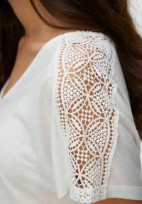Primer plano de la manga de una blusa blanca con detalles intrincados de encaje floral sobre un fondo de cabello largo y castaño y piel.