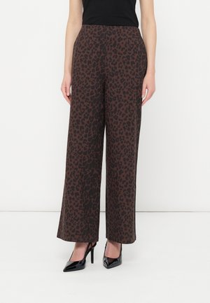 Femme portant un pantalon marron taille haute à jambes larges avec imprimé léopard noir et des talons hauts noirs à bout pointu, debout devant un fond blanc.