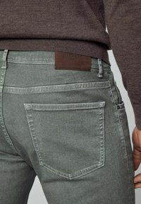 Jean en denim gris avec un design de poche arrière, portant une étiquette en cuir marron embossée de "Hackett London". Texture lisse, coupe ajustée.