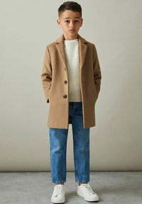 Kamelul frakke med notched lapels, enkel brystkonstruktion, sorte knapper; parret med en lys sweater, blå jeans og hvide sneakers.