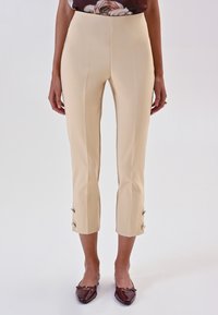 Pantaloni beige su misura con una texture liscia, orlo accorciato e bottoni dorati decorativi ai polsini, abbinati a ballerine marroni.