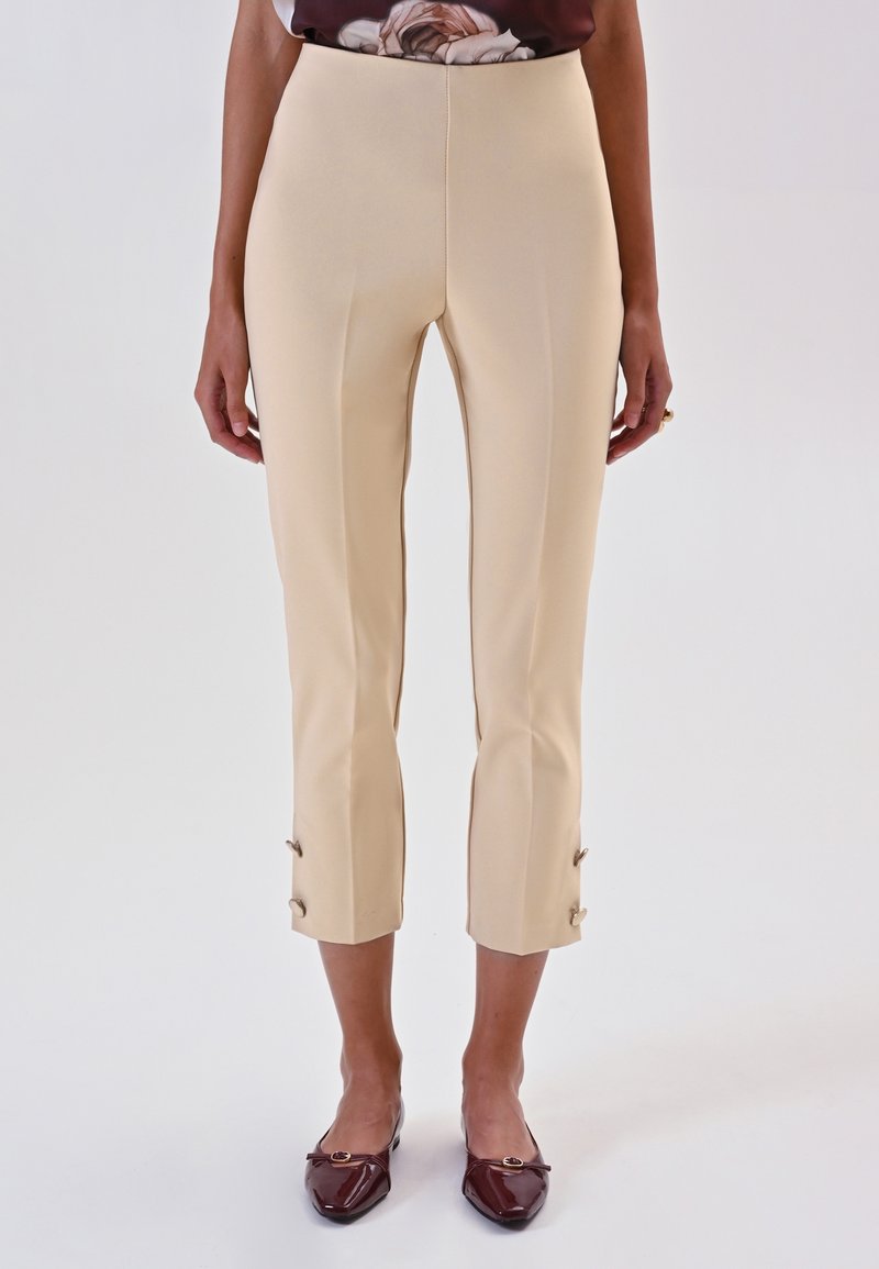 Pantaloni beige su misura con una texture liscia, orlo accorciato e bottoni dorati decorativi ai polsini, abbinati a ballerine marroni.
