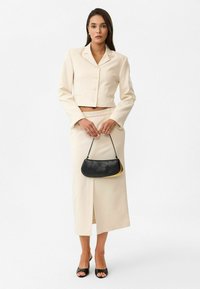 Blazer crème court avec des boutons, jupe taille haute assortie avec une fente, et un petit sac à main noir et jaune. Talons ouverts noirs.