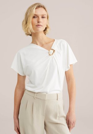 Donna bionda che indossa una maglia bianca a maniche corte con una fibbia dorata vicino allo scollo e pantaloni beige a vita alta con pieghe.