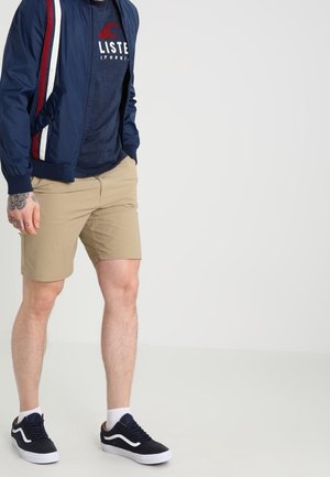 Marine bomberjack met rode en witte zijstrepen, gecombineerd met een marine t-shirt, beige shorts en zwarte sneakers met witte zolen.