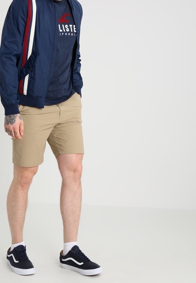 Granatowa kurtka bombowiec z czerwono-białymi paskami po bokach, zestawiona z granatowym podkoszulkiem, beżowymi szortami i czarnymi trampkami z białą podeszwą.