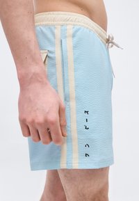 Vue latérale d'une personne portant un short de bain bleu clair avec des rayures beiges et un texte vertical noir sur la cuisse droite.