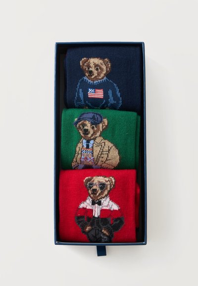Trois chaussettes pliées avec des motifs d'ours en peluche portant différentes tenues, disposées verticalement dans une boîte rectangulaire sombre sur fond blanc.