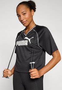 Maillot de sport noir avec des bordures blanches et le logo "PUMA", à manches courtes, coupe décontractée, tenu avec une corde à sauter dans les mains, accompagné de leggings noirs.