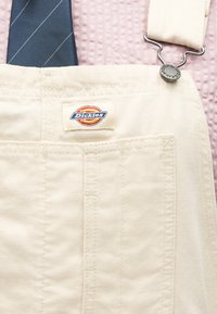 Κρεμ ολόσωμη φόρμα Dickies με μεταλλική αγκράφα και μπροστινή τσέπη στο στήθος, φορεμένη πάνω από ένα ροζ υφασμάτινο πουκάμισο και μια μπλε ναυτική γραβάτα με ρίγες.