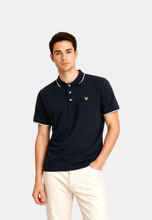 Jeune homme portant un polo bleu marine avec des bordures blanches et un pantalon beige clair, debout avec une main dans la poche sur un fond uni.