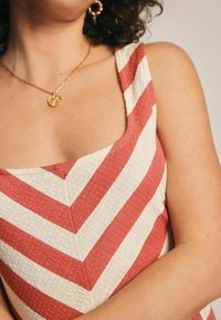 Robe à rayures rouges et blanches avec un tissu texturé, décolleté carré et larges bretelles, accentuée par un collier en pièces dorées.