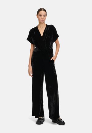 Sort velour jumpsuit med korte ærmer, V-udskæring, bundet talje, vide ben og sidelommer. Båret med kraftige sorte sko.