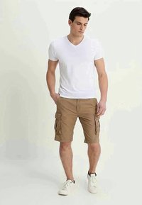 Calções cargo bege com bolsos laterais e cordão, combinados com uma t-shirt branca simples em forma de V. Modelo a usar ténis brancos.