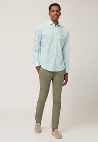 Light blue striped button-up shirt with a small logo, paired with olive green chinos and beige slip-on shoes. Smooth fabric texture.