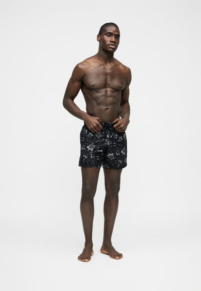 Γυμνόστηθος άντρας όρθιος ξυπόλητος, φοράει μαύρο μαγιό με λευκά μοτίβα χελώνας και φυσαλίδων, προσαρμόζοντας το κορδόνι στη μέση.