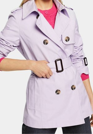 Femme ajustant la ceinture d’un trench-coat croisé violet clair avec de gros boutons, portant un pull rose vif et un pantalon sombre.