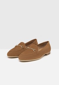 Tan suede loafers featuring a gold chain accent, rounded toe, low heel, and stitched detailing along the edges. Smooth inner lining.