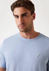 Joven con cabello castaño claro y barba, vistiendo una camiseta azul claro con un pequeño logo triangular de Guess en el pecho.