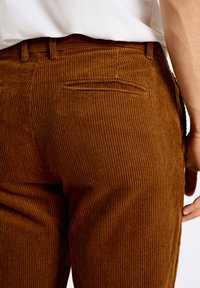 Pantalons en velours côtelé marron avec un ribbing vertical, dotés d'une poche arrière et de passants de ceinture, associés à une chemise blanche unie.