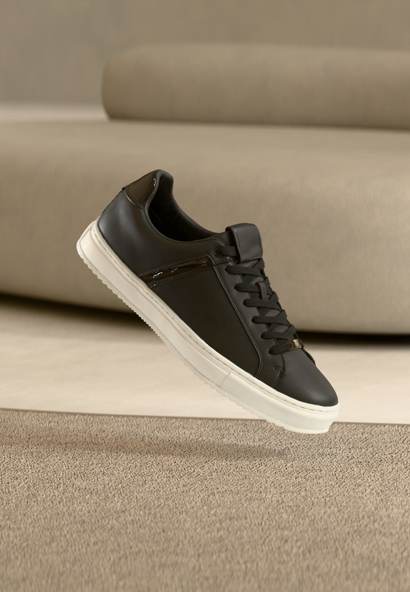 Baskets en cuir noir avec semelle blanche et lacets noirs, suspendues au-dessus d'un tapis beige avec un canapé beige en arrière-plan.