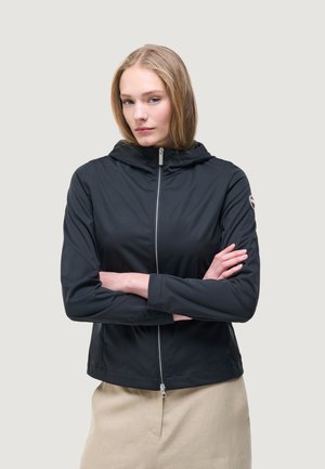 Žena se světle hnědými vlasy, oblečená v černé zipové bundě s kapucí, se založenýma rukama a béžovou sukní, na jednoduchém světlém pozadí.