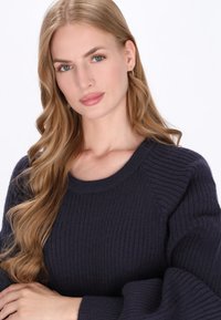 Mørk navy ribbestrikket sweater med rund hals, der har tekstureret stof og lange, let puffede ærmer. Ingen synlige mønstre eller hardware.