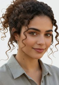 Porträtt av en person med lockigt brunt hår, iklädd en ljusgrå skjorta med krage och silverfärgade hoops, mot en enfärgad bakgrund.