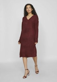 Robe en tricot bordeaux avec un décolleté en V et des manches longues, dotée de fentes latérales à l'ourlet. Associée à des sandales à talons hauts bordeaux à lanières.