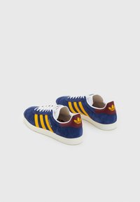 Blå mockasneakers med tre gula ränder, vita skosnören och en vinröd hälflik med en logotyp. Klassisk lågskuren design.