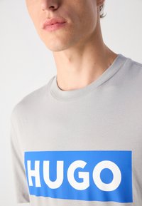 Pilkas trumpas rankovėmis marškinėliai su mėlynu stačiakampiu grafiniu pavaizdavimu, kuriame dideliu baltu tekstu užrašyta "HUGO" krūtinėje. Minkštas audinio tekstūros.