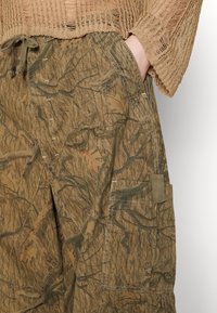 Cargo-byxor i kamouflage-mönster med nyanser av brunt och grönt, tillverkade av texturerad tyg med synlig sömnad och en sidofickdetalj.