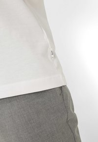 Maglietta bianca in cotone con etichetta laterale, abbinata a pantaloni grigi chiari e strutturati. La maglietta presenta un design semplice e pulito.