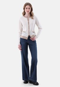 Beige Strickjacke mit rundem Ausschnitt, vorderen Knöpfen und zwei Taschen, kombiniert mit dunkelblauen weit geschnittenen Jeans und schwarzen Plateauschuhen.