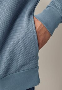 Sudadera azul claro con tejido acolchado texturizado, que presenta un puño acanalado y un detalle de bolsillo. El material parece suave y cómodo.