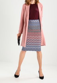 Abrigo texturizado rosa, blusa burdeos, falda lápiz con estampado de chevron en azul y rojo, bolso de mano negro, tacones negros.