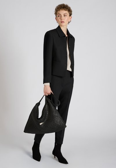 Czarna pleciona torba tote o trójkątnym kształcie i gładkiej fakturze, trzymana przez osobę ubraną w czarną kurtkę i dopasowane spodnie.