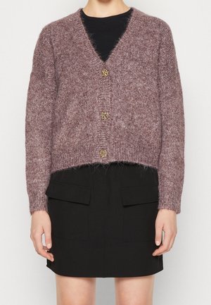 Kobieta ubrana w krótki, puszysty kardigan w kolorze szarofioletowym z trzema guzikami w panterkę, na czarnej bluzce i czarnej spódnicy z przednimi kieszeniami.