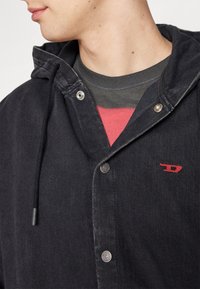 Ung person iklädd en svart jacka med huva och silverfärgade tryckknappar, samt en röd broderad logotyp på bröstet, över en grå och röd t-shirt.