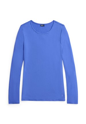 Femme aux cheveux bouclés portant une chemise bleue à manches longues et un short vert à rayures, les mains dans les poches, sur un fond uni.