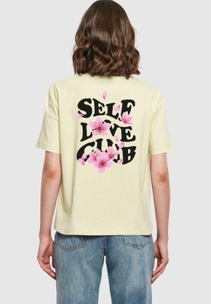 Gelbes T-Shirt mit kurzen Ärmeln, das den Text "SELF LOVE CLUB" in Schwarz zeigt, verziert mit pinken Blumen auf der Rückseite.