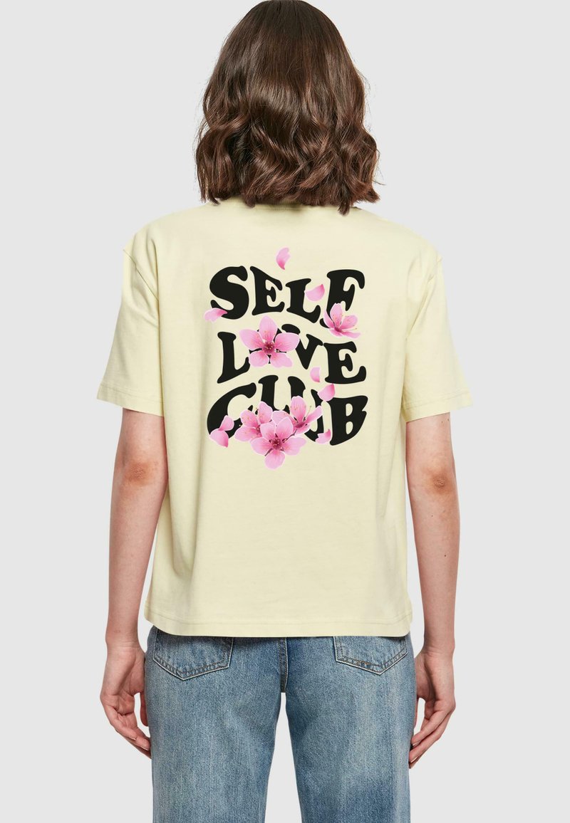 Žluté tričko s krátkým rukávem, na kterém je text "SELF LOVE CLUB" v černé barvě, doplněný růžovými květinami na zádech.