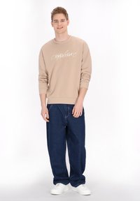 Beige pulóver "endless" felirattal, laza szabású sötétkék farmerrel párosítva. A modellen fehér sportcipő van, egy egyszínű háttér előtt áll.