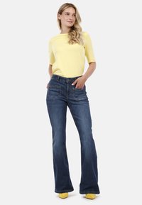 Top corto de color amarillo claro con mangas cortas combinado con jeans de campana azul oscuro que presentan dos bolsillos frontales. Unos zapatos amarillos complementan el atuendo.