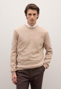 Maglione beige in lana con scollo rotondo, polsini e orlo a coste, indossato sopra una camicia bianca con colletto, abbinato a pantaloni marrone scuro.