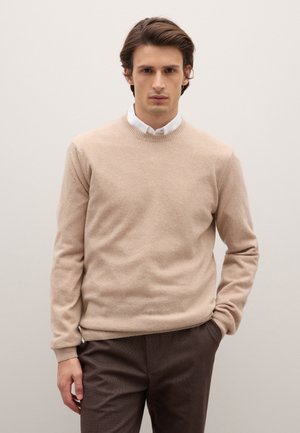 Maglione beige in lana con scollo rotondo, polsini e orlo a coste, indossato sopra una camicia bianca con colletto, abbinato a pantaloni marrone scuro.